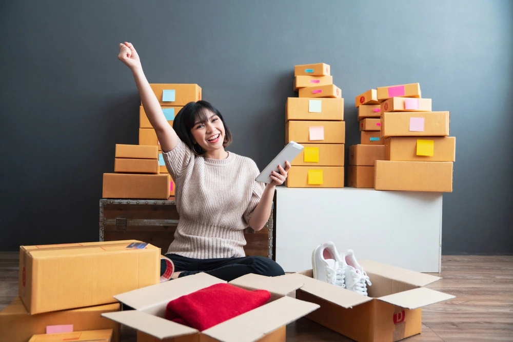 Woman happy as she's packing boxes of clothes and shoes with tablet in hand.