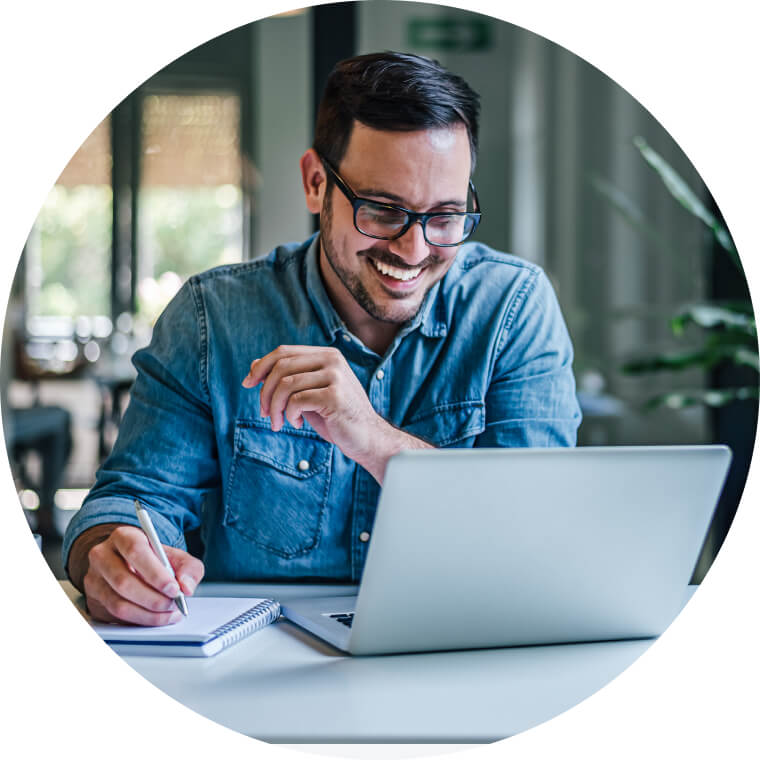 An  a man smiling whilst using a pen and notepad infront of a laptop.