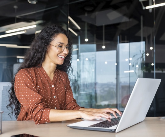 A woman sat at a desk working on a laptop smiling.