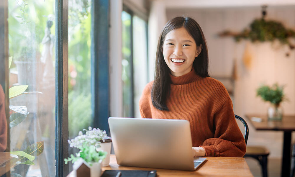 Woman smiling while seated at table using a laptop, ready for QuickBooks On-Demand Webinar Training