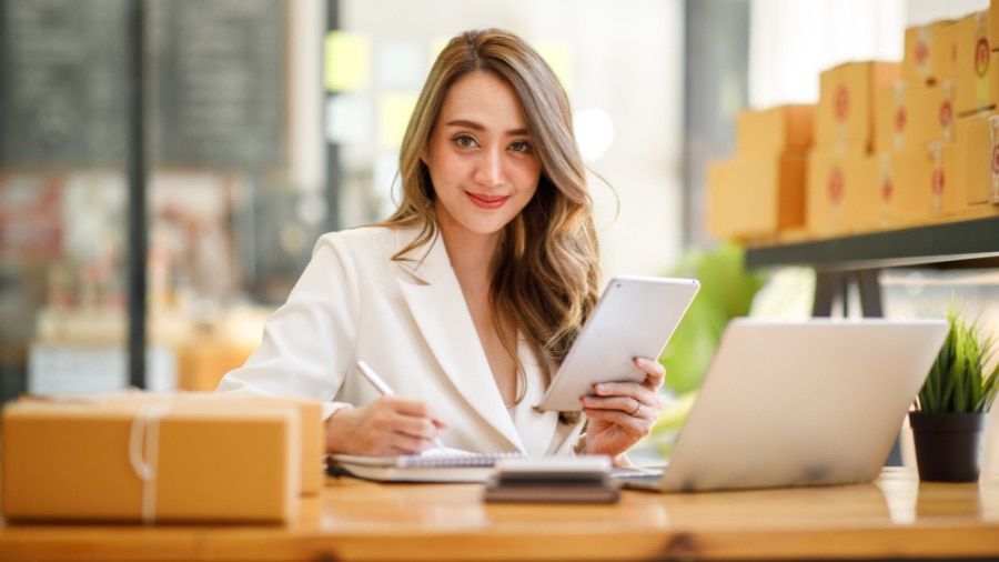 A woman looking at the camera with a notepad in hand and a laptop on the table 