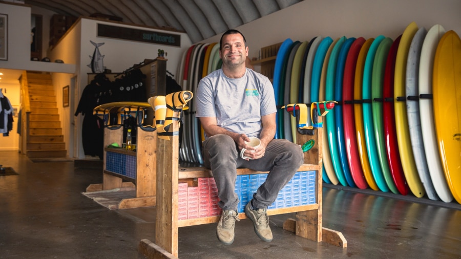 A person sitting on a wooden bench with a bunch of surfboards.
