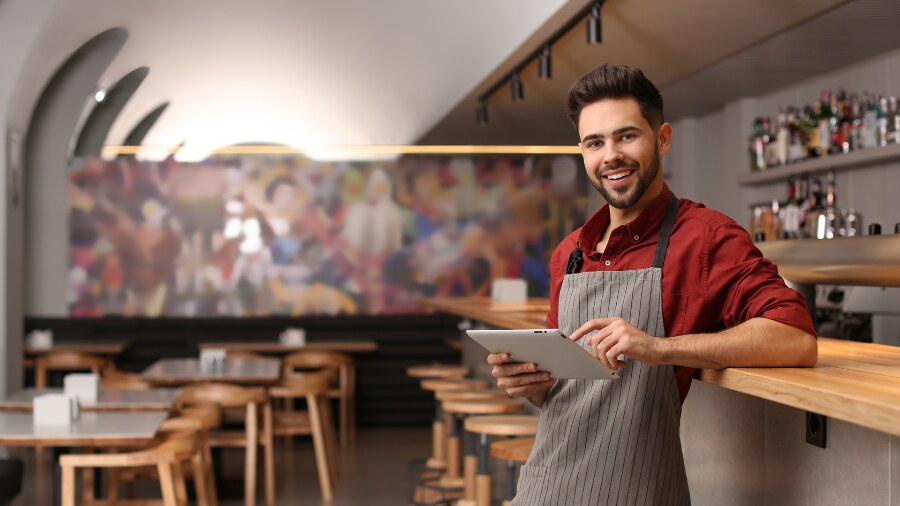 A male business owner standing in front of a table holding an electronic tablet. 