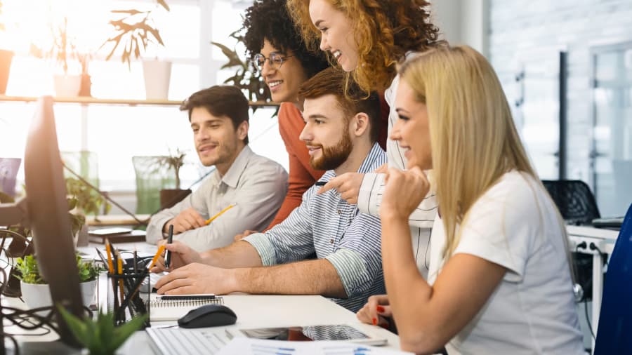 A marketing team stands over a worker and smiles at the results they see on the monitor.