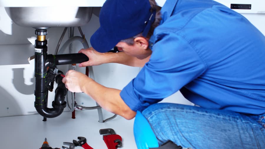 A plumber crouches beneath a sink and fixes the pipes with a wrench. 