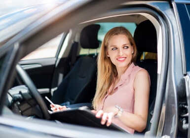 A smiling woman holds a tablet while sitting in a parked car.