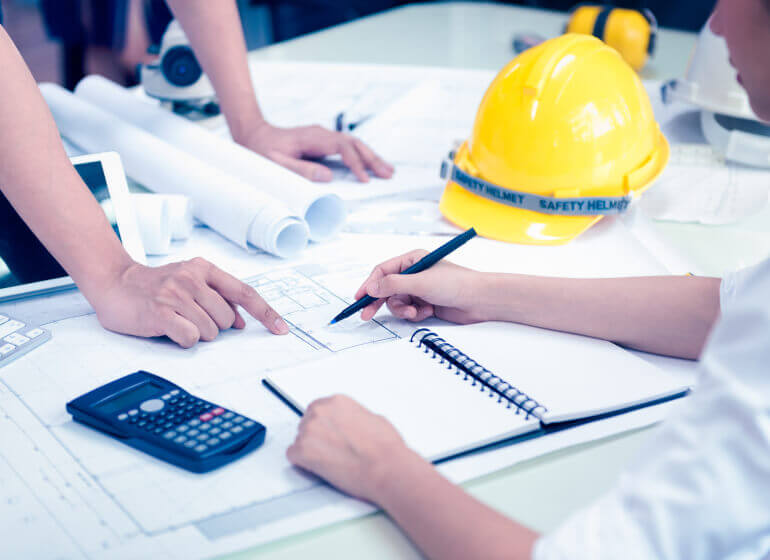 The hands of two workers reach over a table with a notebook, calculator, construction hat and other projected paperwork.