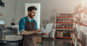 A man stands in front of coffee bean shelf, and holding a laptop.