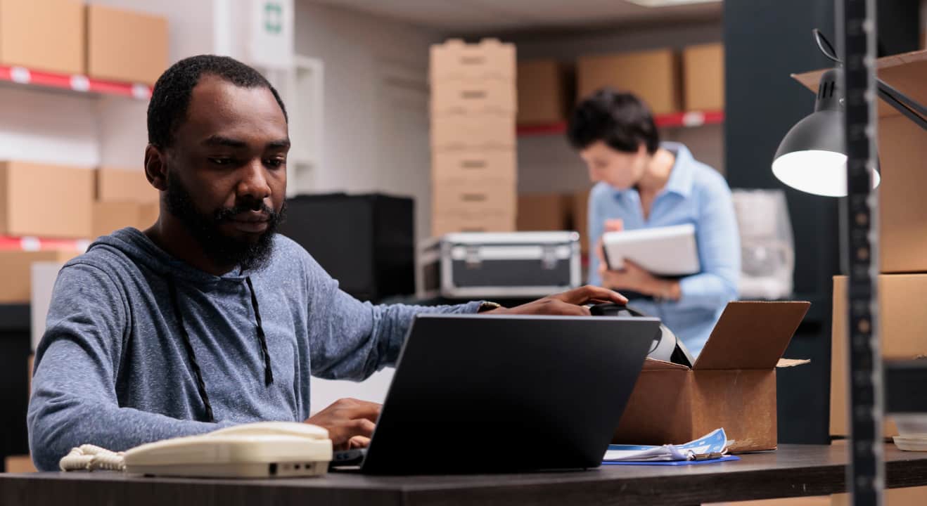 A manager in a stock room concentrates on his laptop at his desk, while workers check inventory in the background.