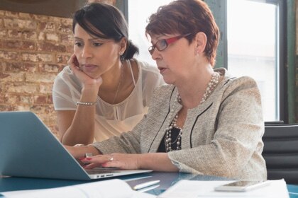 Two well dressed women confer while examining a laptop display.