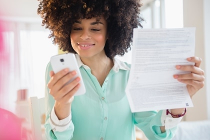 A woman in a turquoise shirt holds up her phone and a paper form and smiles.