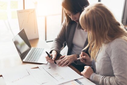 Two business women check details on a print out beside their laptop.