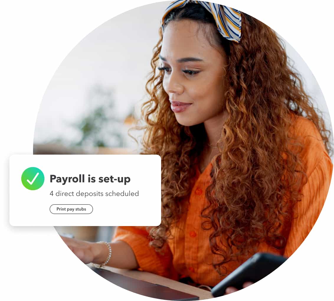 A woman with curly hair wearing an orange shirt works on her laptop with her phone showing payroll is set up in QuickBooks