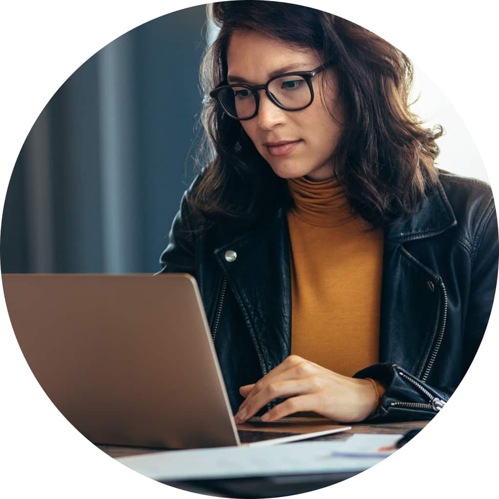 A woman wearing glasses and a leather jacket focuses intently on her laptop computer.