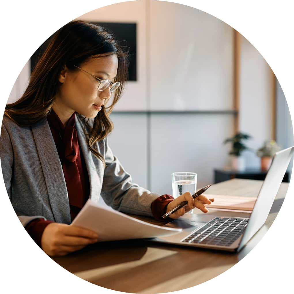 A sharply dressed woman wearing a blazer types on her laptop while referencing documents.