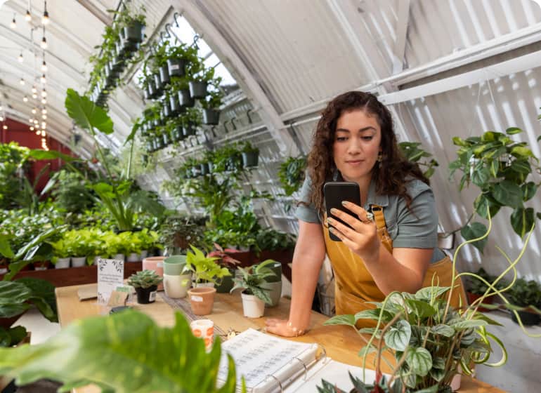 A business owner looks at her smartphone inside a greenhouse full of plants.