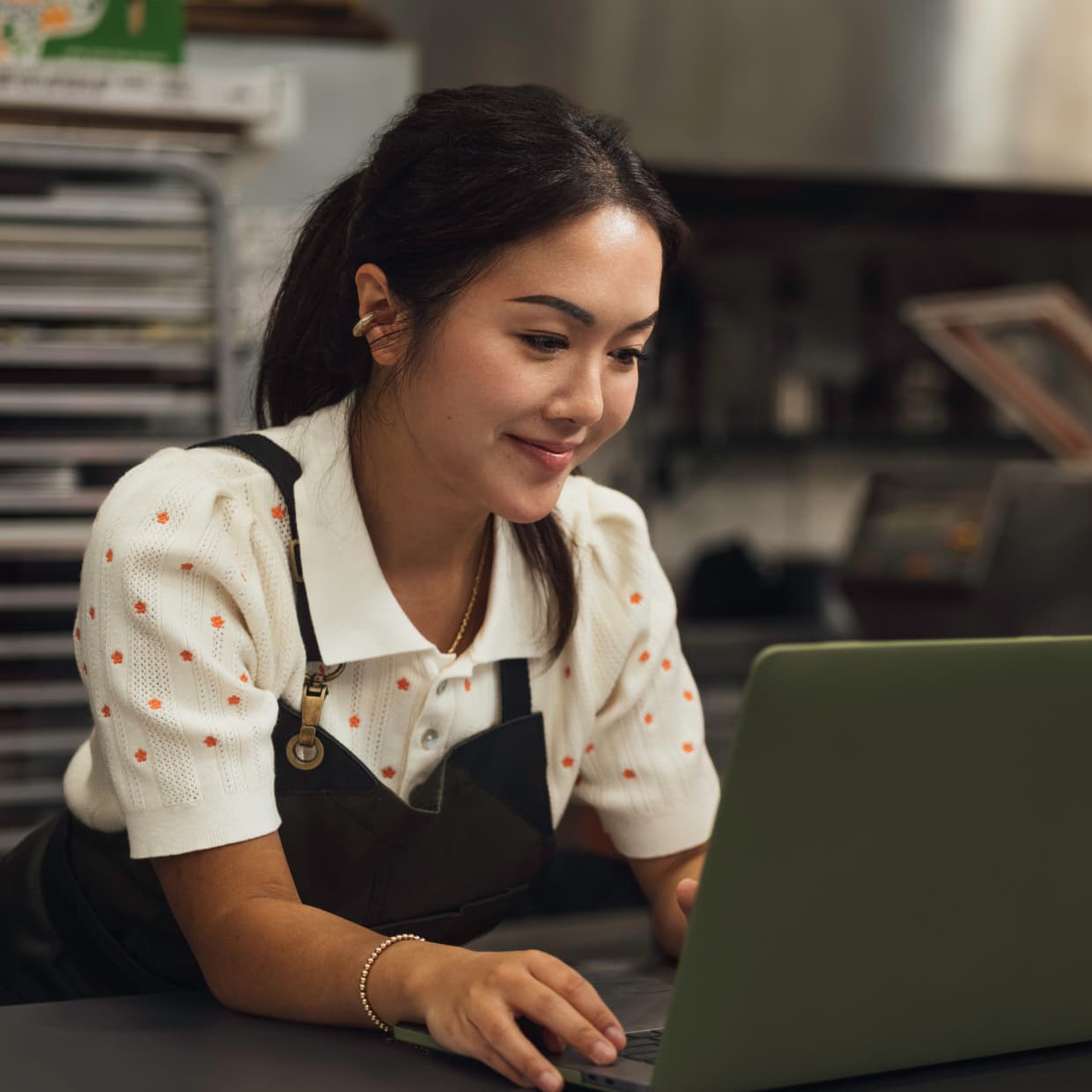 A woman wearing an apron smiles as she works on her laptop in a clean restaurant kitchen.