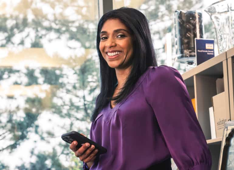 Owner of Sahajan Beauty leans against her desk and smiles while holding her cell phone.