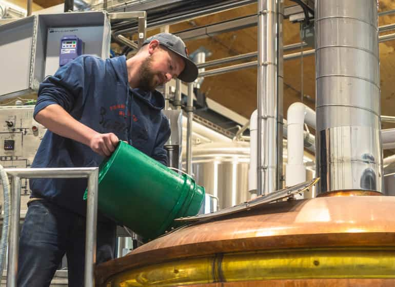 A brewery worker pours from a green can into a large metal vat.