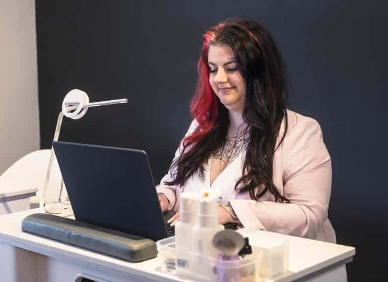 The owner of Pride Beauty Lounge sits at a counter with beauty supplies while working on her laptop. 