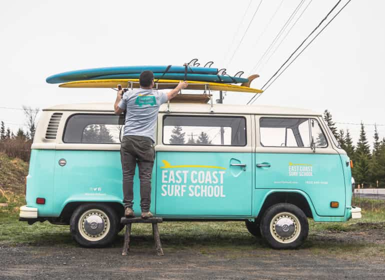 The founder of East Coast Surf School stands on a stool to reach his surf boards that are mounted on top his teal VW van.