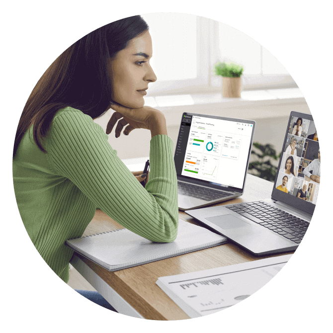 A woman engages in an online meeting on a laptop. A second laptop on her desk shows a QuickBooks Online dashboard.