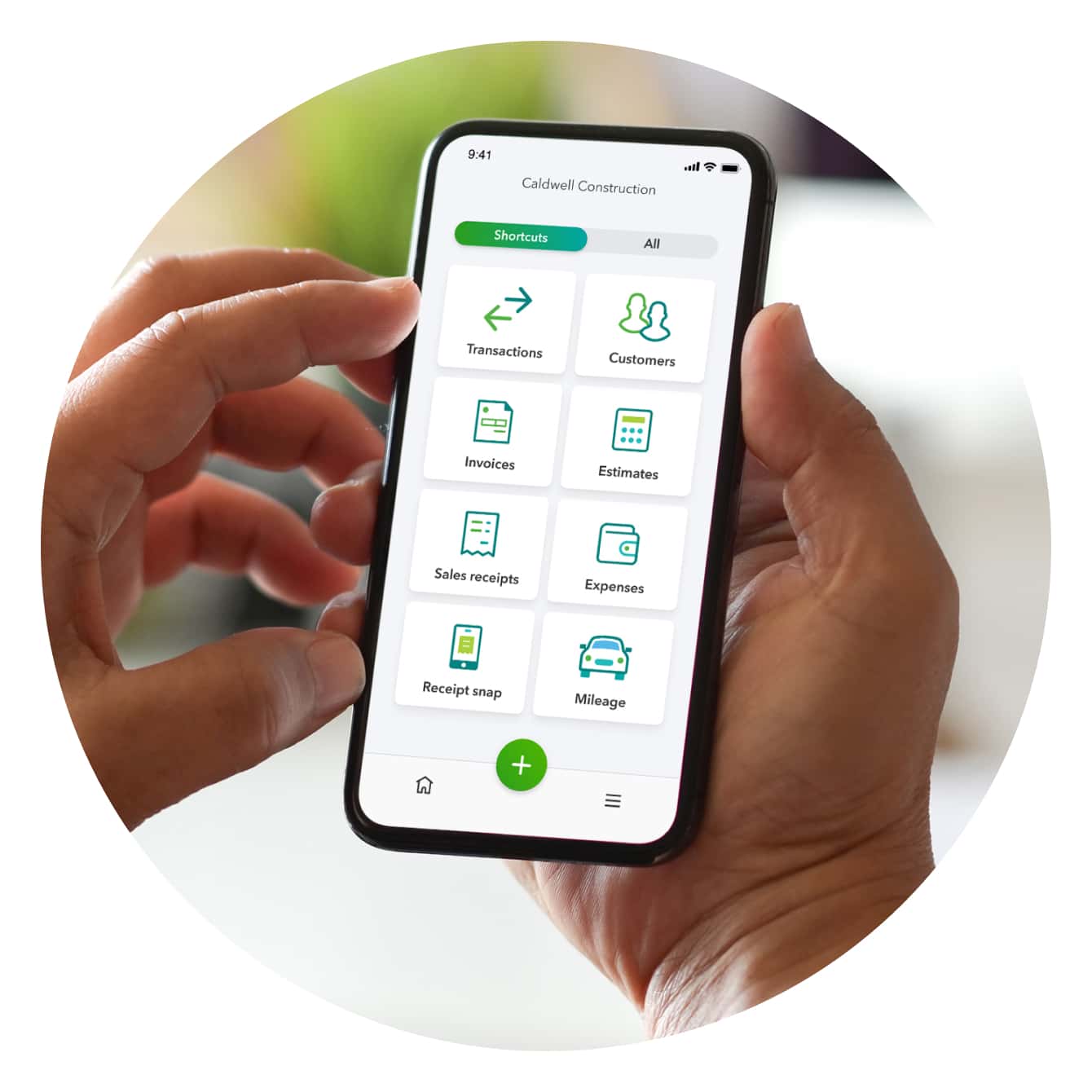 A hand holding a smartphone showing QuickBooks Online shortcuts – transactions, customers, invoices, estimates and more.