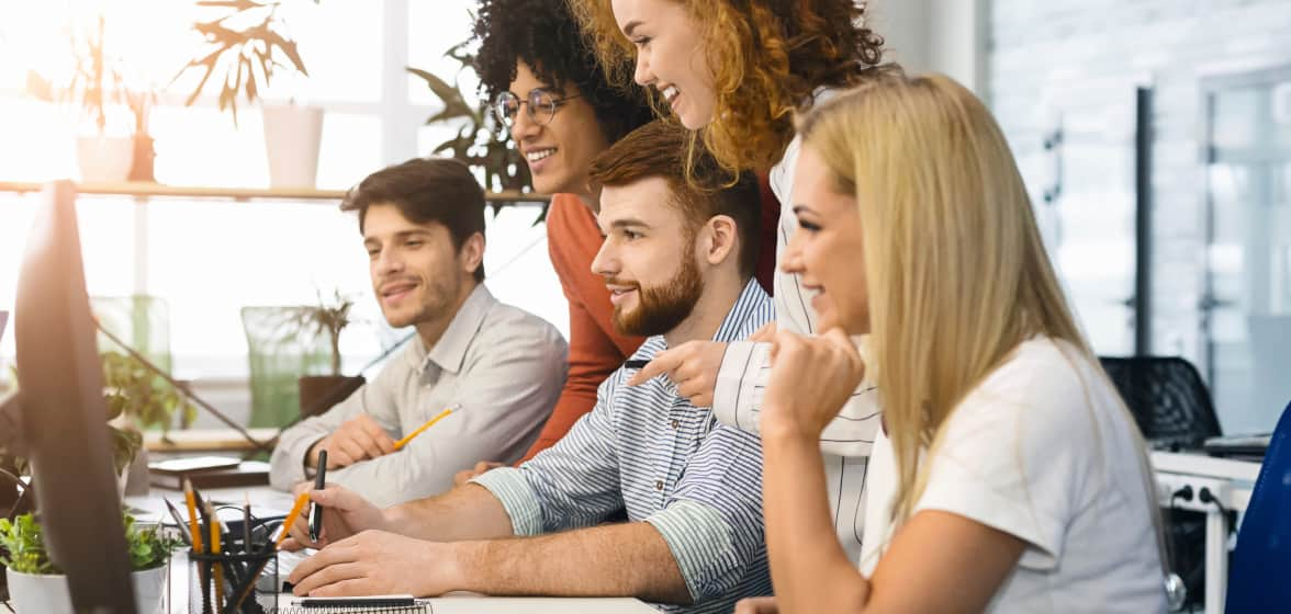 A marketing team stands over a worker and smiles at the results they see on the monitor.