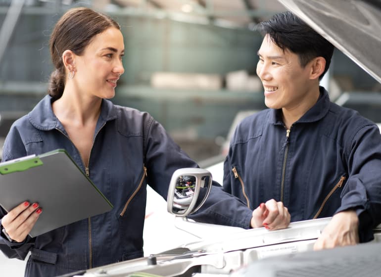 Two mechanics focus over the engine of a car beneath the raised front hood.