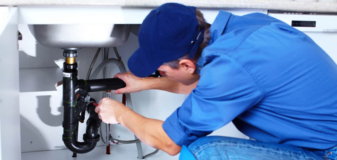 A plumber crouches beneath a sink and fixes the pipes with a wrench. 