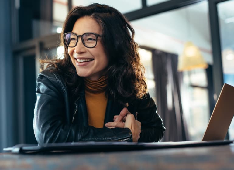 A woman wearing a leather jacket smiles as she looks away from her sleek laptop computer. 