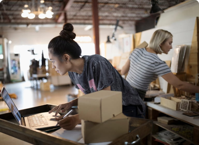 A woman works on her laptop while her colleague works in the background of their workshop.