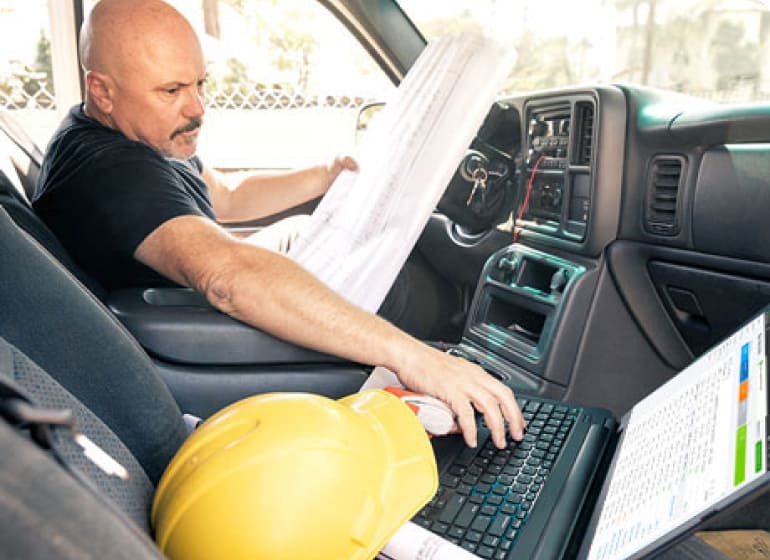 A construction worker works on his laptop while sitting in a parked car. His construction hat rests next to the laptop.