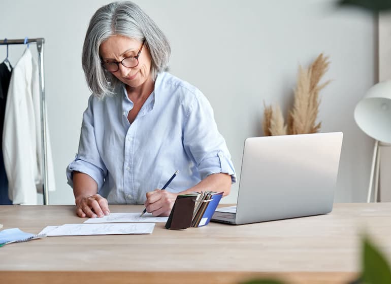 Une femme âgée est assise à un bureau et écrit, avec son ordinateur portable à ses côtés.