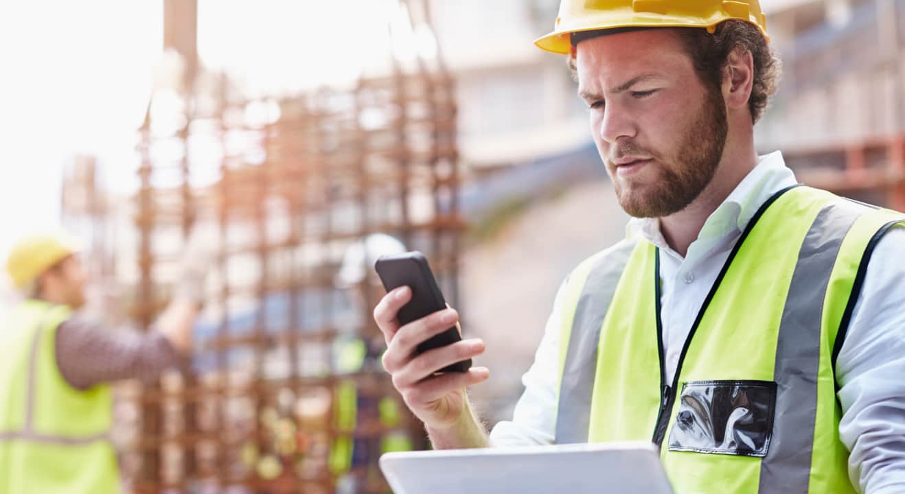 A bearded construction manager checks his phone and tablet while at a work site.