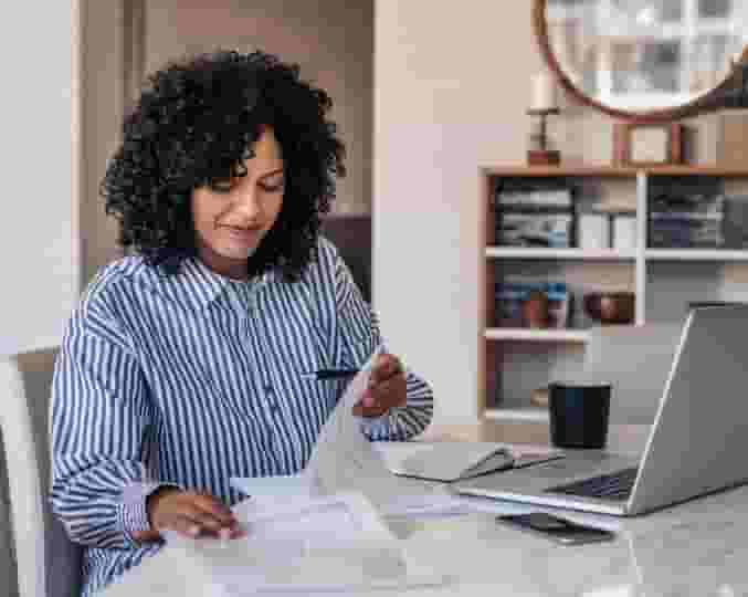 Une femme portant une chemise à rayures lit des documents devant son ordinateur portable