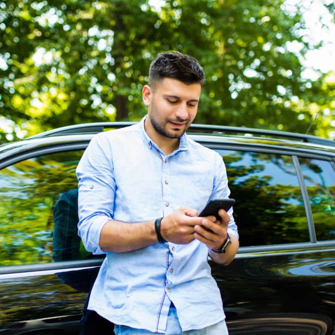 A man leans back against his SUV while consulting the QuickBooks Mobile App on his phone.