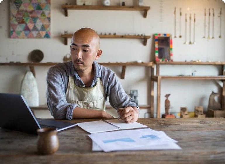 A man in a workshop writes in a notebook while glancing at his Apple laptop.