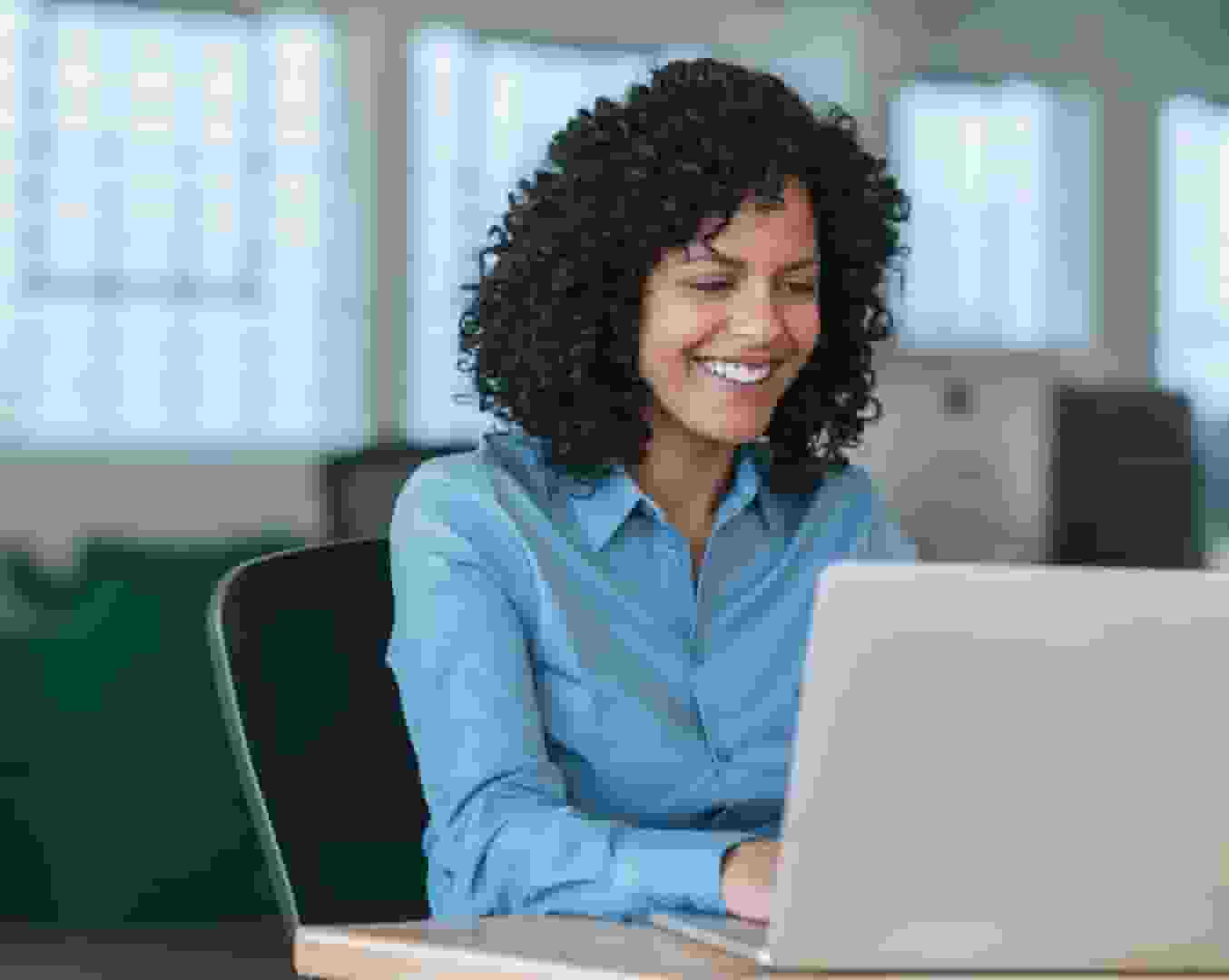 Une femme aux cheveux noirs bouclés portant une chemise bleue sourit en travaillant sur son ordinateur portable