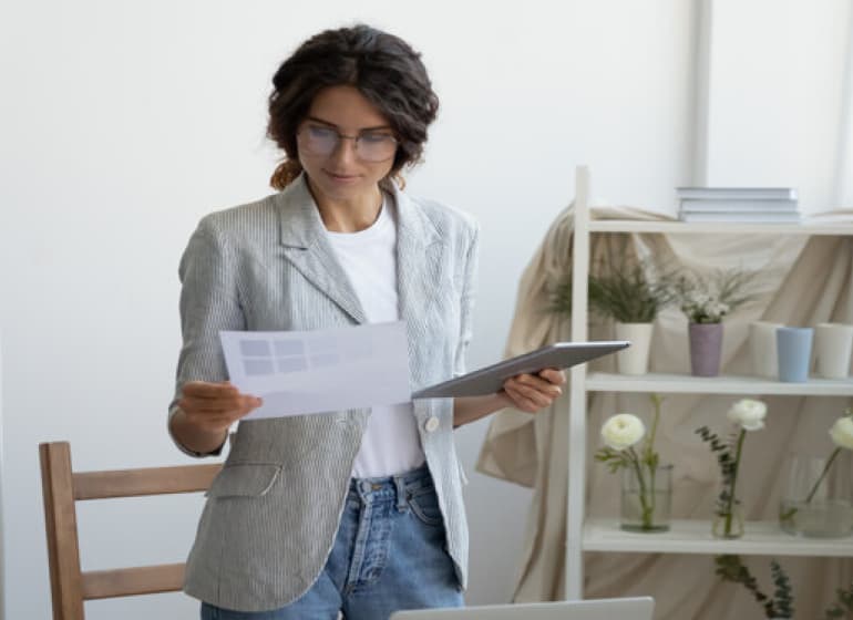 Une femme portant un blazer gris examine une impression dans une main et tient sa tablette informatique dans l'autre.