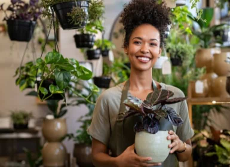 A woman holds a potted plant in her shop and smiles warmly. 