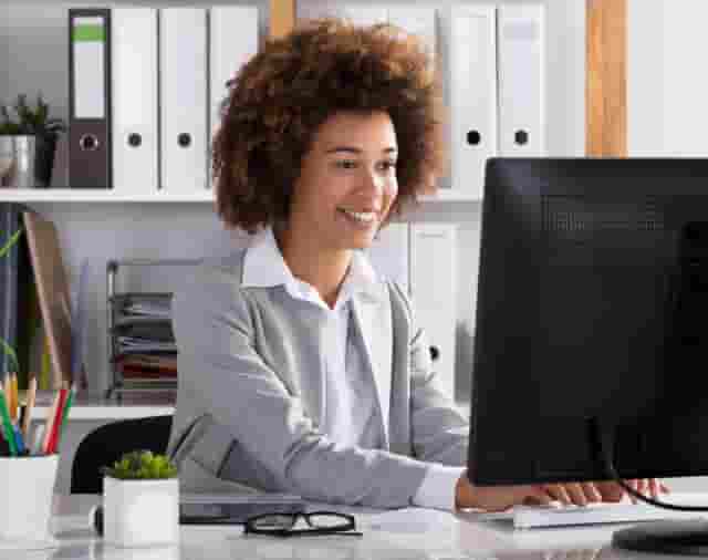 Femme souriante en pull gris travaillant sur ordinateur avec un grand écran gris foncé.