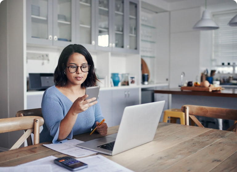 A woman checks her smartphone while working at laptop at her kitchen table.