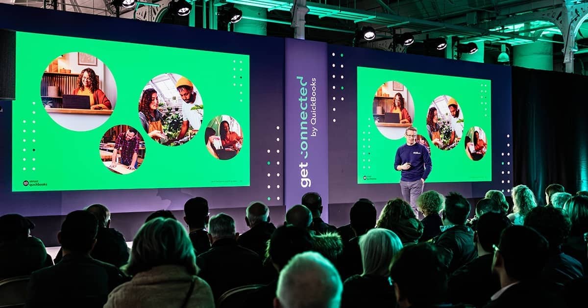 A group of people sitting at a podium with a screen on it.