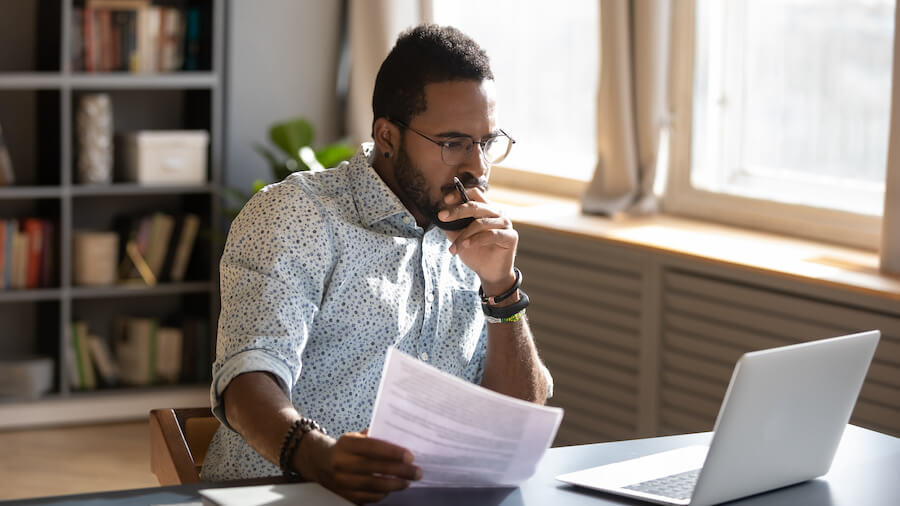 Man sitting at desk holding documents and reading from a laptop