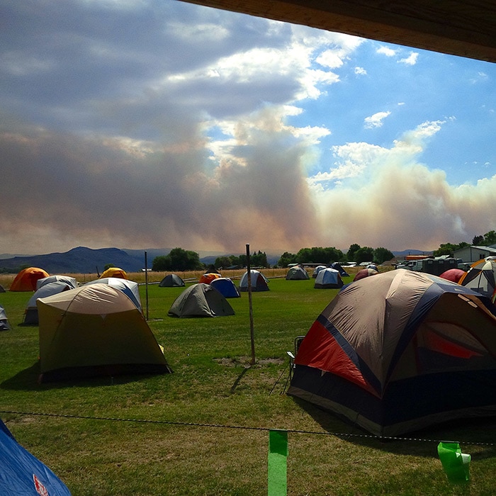 Smoke from a forest fire in Oregon is depicted in the distance beyond the encampment of firefighters, mobile kitchen staff, and other supporting personnel.