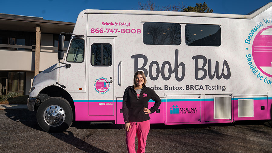 A woman in a pink and black fleece stands proudly in front of The Boob Bus, a large mobile mammography unit painted white with bright pink branding.