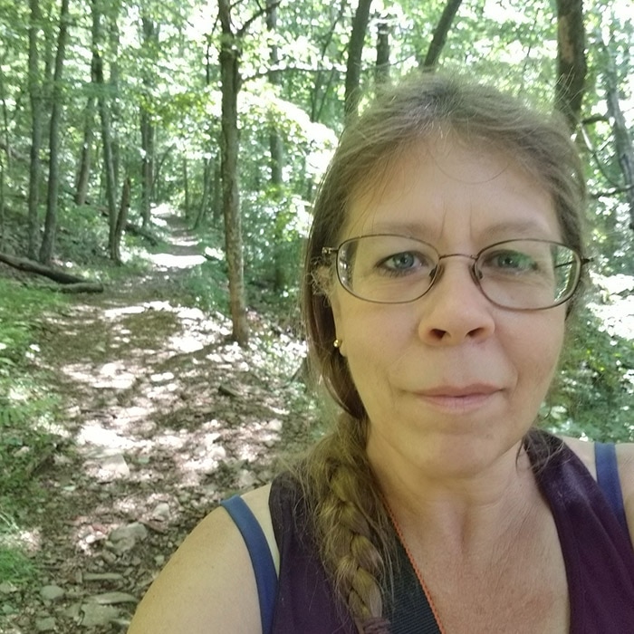 A person in a green shirt and jeans taking a selfie in a forest.