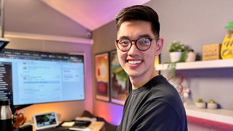 A smiling person in glasses and a black shirt in front of a computer.