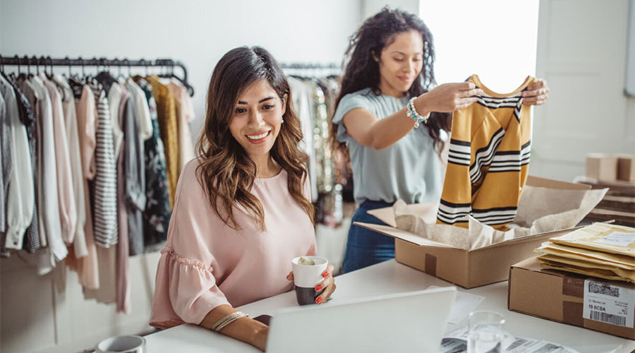 Two business partners manage day-to-day operations in a clothing store. The person on the left has brown hair, wears a pink shirt, and is looking at a computer. The woman on the right has curly hair and is folding up a striped shirt to ship to a customer.